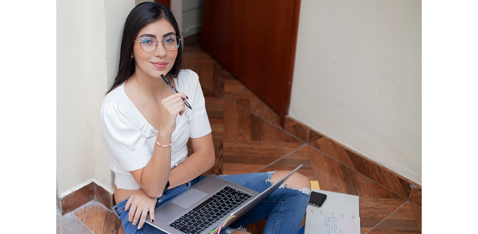 College age Latin woman with long hair, wears reading glasses. She is sitting on the floor of her dorm with the laptop on her legs while looking at the camera and smiling.