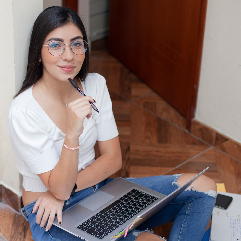College age Latin woman with long hair, wears reading glasses. She is sitting on the floor of her dorm with the laptop on her legs while looking at the camera and smiling.